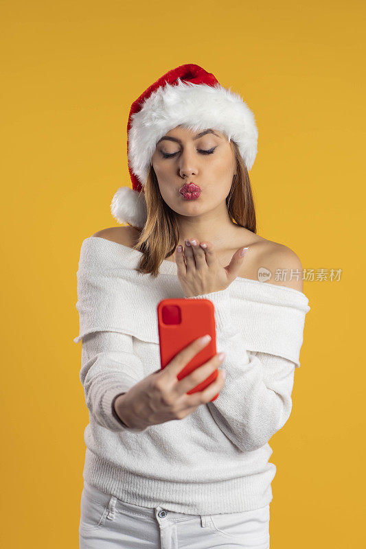 美丽的年轻女子戴着圣诞老人的帽子，对着她的智能手机相机飞一个吻，在黄色背景上进行视频通话/自拍